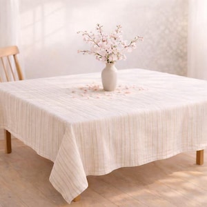 May include: A square, cream-colored tablecloth with vertical stripes covers a wooden table. A white vase holds a bouquet of pink flowers. The table is set in a well-lit room, suggesting a dining or living space.