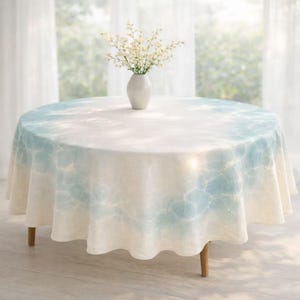 May include: A round tablecloth with a light blue and white water-like pattern. The tablecloth is draped over a wooden table. A white vase with white flowers sits in the center of the table.