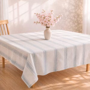 May include: A white tablecloth with blue stripes covers a wooden table. A white vase holds pink flowers. The tablecloth has a classic, farmhouse style, suitable for dining or decorative use.