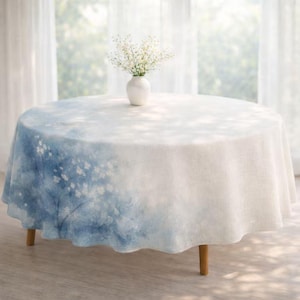 May include: A round table with a white tablecloth featuring a blue watercolor floral design. A small white vase with white flowers sits on the table. The table has wooden legs and is set in front of a window.