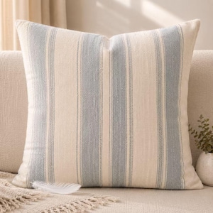 May include: Square throw pillow with a cream-colored background and vertical light blue stripes. The pillow is made of a textured fabric and is placed on a neutral-colored sofa. A white feather rests on the sofa.