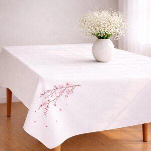 May include: A white rectangular tablecloth with a delicate pink cherry blossom branch design in the corner. A white vase with white flowers sits on the table. The table has wooden legs.