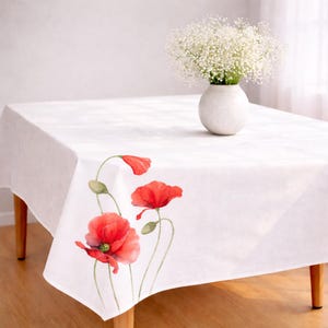 May include: White rectangular tablecloth with a watercolor design of red poppies and green stems. A small white vase holds a bouquet of white flowers. The tablecloth is on a wooden table.