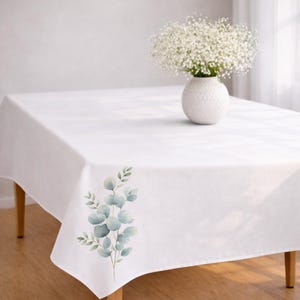 May include: A white rectangular tablecloth with a watercolor eucalyptus design in shades of green and blue on one corner. A white vase with a bouquet of white flowers sits on the table. The table has wooden legs.