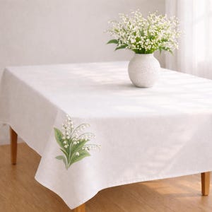 May include: A white rectangular tablecloth with a floral design in the corner. A white vase with a bouquet of white flowers sits on the table. The table has wooden legs and is set on a wooden floor.