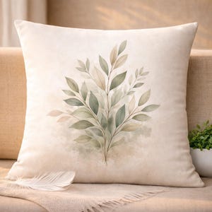 May include: A square, cream-colored decorative pillow featuring a watercolor design of green and tan leaves. The leaves are arranged in a symmetrical pattern against a soft, neutral background. A white feather rests on the sofa.