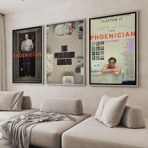 Könnte beinhalten: Drei gerahmte Filmplakate mit dem Text "THE PHOENICIAN SCHEME" in Rot und Lila. Die Poster hängen über einem hellgrauen Ecksofa mit Kissen und einer Decke. Die Poster haben schwarze Rahmen.