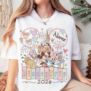 K&ouml;nnte beinhalten: Wei&szlig;es T-Shirt mit einem Disney-Motiv. Das Design zeigt Micky und Minnie Maus, Donald Duck und andere Charaktere, zusammen mit dem Text "DisneyTrip 2026". Die Grafik ist von Sternen und Ballons umgeben.
