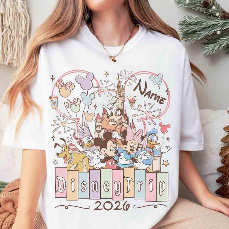 K&ouml;nnte beinhalten: Wei&szlig;es T-Shirt mit einem Disney-Motiv. Das Design zeigt Micky und Minnie Maus, Donald Duck und andere Charaktere, zusammen mit dem Text "DisneyTrip 2026". Die Grafik ist von Sternen und Ballons umgeben.
