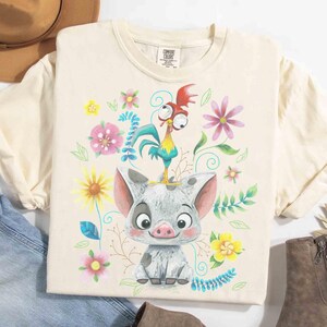May include: Cream-coloured t-shirt featuring a cartoon pig with a rooster perched on its head, surrounded by colourful flowers and green leaves. The shirt has a Comfort Colors tag.