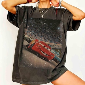 Puede incluir: Camiseta gris oscuro con un gr&aacute;fico del personaje de Cars, Rayo McQueen. La imagen muestra a Rayo McQueen corriendo en una pista con otros coches y una multitud de fondo. La camiseta tiene un aspecto vintage y desgastado.