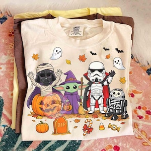 May include: A cream-colored t-shirt featuring a graphic of Star Wars characters in Halloween costumes, including a mummy Darth Vader in a pumpkin, a witch Grogu, a vampire Stormtrooper, and a skeleton R2-D2, with ghosts, bats, and leaves.