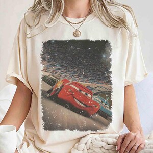 Puede incluir: Camiseta color crema con un gr&aacute;fico del personaje animado Rayo McQueen de la pel&iacute;cula Cars. La imagen muestra el coche de carreras rojo en acci&oacute;n en un circuito, con un fondo de cielo estrellado.