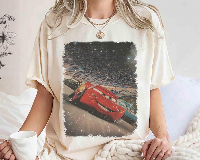 Puede incluir: Camiseta color crema con un gr&aacute;fico del personaje animado Rayo McQueen de la pel&iacute;cula Cars. La imagen muestra el coche de carreras rojo en acci&oacute;n en un circuito, con un fondo de cielo estrellado.