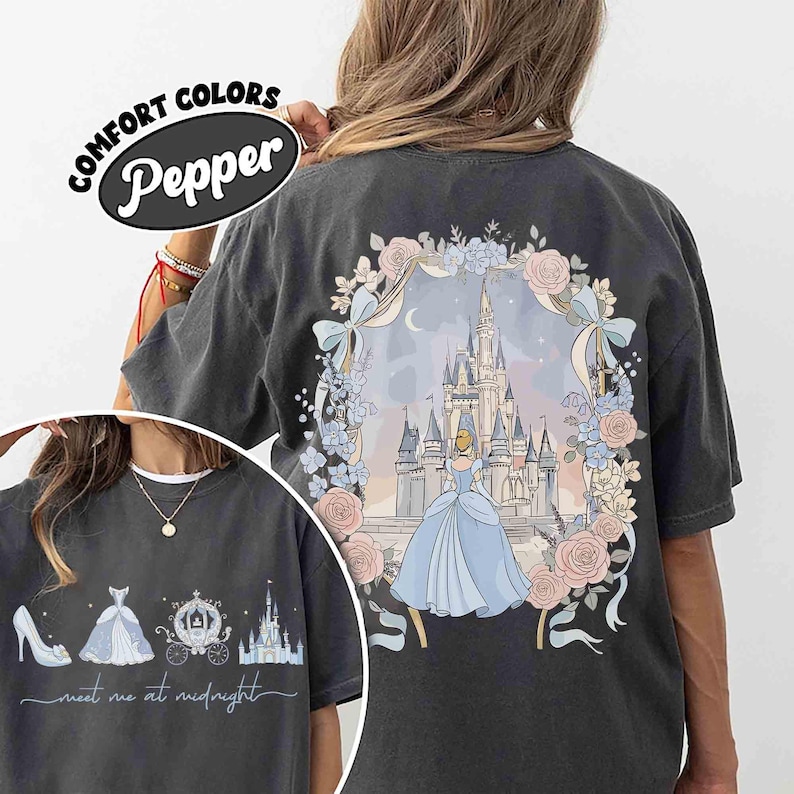 May include: Dark gray t-shirt featuring a Cinderella design. The back of the shirt has a castle illustration with Cinderella in a blue gown, surrounded by flowers. The front has a shoe, dress, carriage, and castle with the text "meet me at midnight".