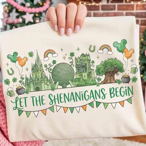 Puede incluir: Camiseta de color crema con un gráfico verde y naranja que representa un castillo, una gran esfera y un árbol. El texto "LET THE SHENANIGANS BEGIN" está escrito en verde. El diseño incluye arcoíris, globos y tréboles.