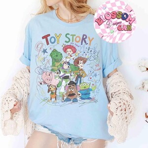 Puede incluir: Camiseta azul claro con un gr&aacute;fico colorido de los personajes de Toy Story, incluyendo a Woody, Buzz Lightyear y Rex. Las palabras "Toy Story" est&aacute;n escritas en una fuente juguetona sobre los personajes. La camiseta tiene las mangas enrolladas.
