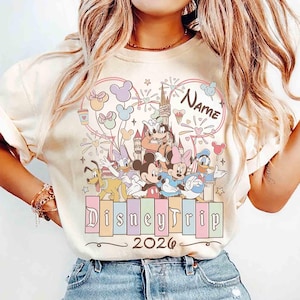 K&ouml;nnte beinhalten: Cremefarbenes T-Shirt mit einem farbenfrohen Disney-Motiv. Das Design zeigt Micky Maus, Minnie Maus, Donald Duck und andere Charaktere, zusammen mit dem Text "DisneyTrip 2026" und Platz f&uuml;r einen Namen. Das Motiv ist von einem herzf&ouml;rmigen Rahmen umgeben.