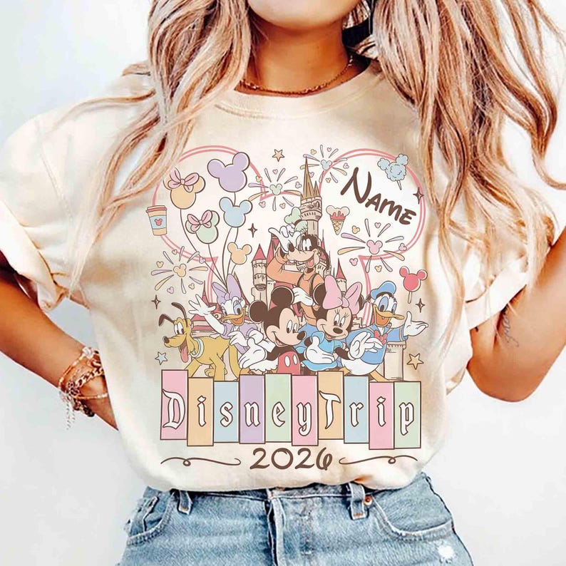 K&ouml;nnte beinhalten: Cremefarbenes T-Shirt mit einem farbenfrohen Disney-Motiv. Das Design zeigt Micky Maus, Minnie Maus, Donald Duck und andere Charaktere, zusammen mit dem Text "DisneyTrip 2026" und Platz f&uuml;r einen Namen. Das Motiv ist von einem herzf&ouml;rmigen Rahmen umgeben.