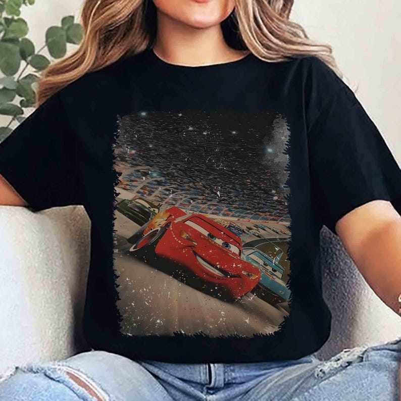Puede incluir: Camiseta negra con un gr&aacute;fico de Rayo McQueen de la pel&iacute;cula Cars corriendo en una pista. La imagen tiene un aspecto vintage y desgastado con un fondo de cielo estrellado. El dise&ntilde;o est&aacute; centrado en la camiseta.