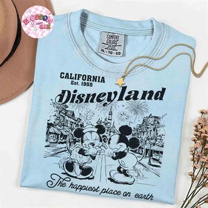 May include: Light blue Disneyland t-shirt with a graphic of Mickey and Minnie Mouse. The shirt features the text "CALIFORNIA Est. 1955 Disneyland" and "The happiest place on earth". A gold star pendant necklace is draped across the shirt.