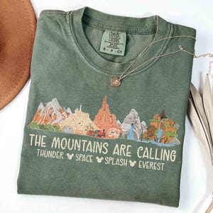 以下が含まれることがあります： オリーブグリーンのクルーネックTシャツ。様々な山の頂上のグラフィックと「THE MOUNTAINS ARE CALLING」というテキスト、その後に「THUNDER」「SPACE」「SPLASH」「EVEREST」という単語が並び、各単語の間に小さなネズミの耳のアイコンが配置されています。
