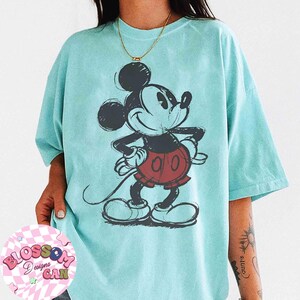 Op de afbeelding: Lichtblauw T-shirt met een geschetste Mickey Mouse-tekening in zwart, rood en wit. De cartoonmuis lacht met de handen op de heupen. Het shirt heeft korte mouwen en een losse pasvorm.