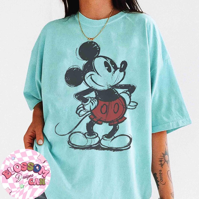 Op de afbeelding: Lichtblauw T-shirt met een geschetste Mickey Mouse-tekening in zwart, rood en wit. De cartoonmuis lacht met de handen op de heupen. Het shirt heeft korte mouwen en een losse pasvorm.