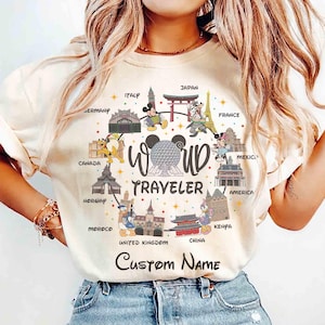 May include: A cream-coloured t-shirt with a world travel design. The shirt displays Mickey and Minnie Mouse in front of global landmarks, with the words "World Traveler" and space for a custom name.