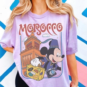 Pode incluir: Camiseta lil&aacute;s clara com um desenho do Mickey Mouse em um traje roxo e azul, segurando uma mala e uma c&acirc;mera. A palavra "Morocco" est&aacute; arqueada sobre uma ilustra&ccedil;&atilde;o de um edif&iacute;cio. O design &eacute; emoldurado em um ret&acirc;ngulo azul claro.