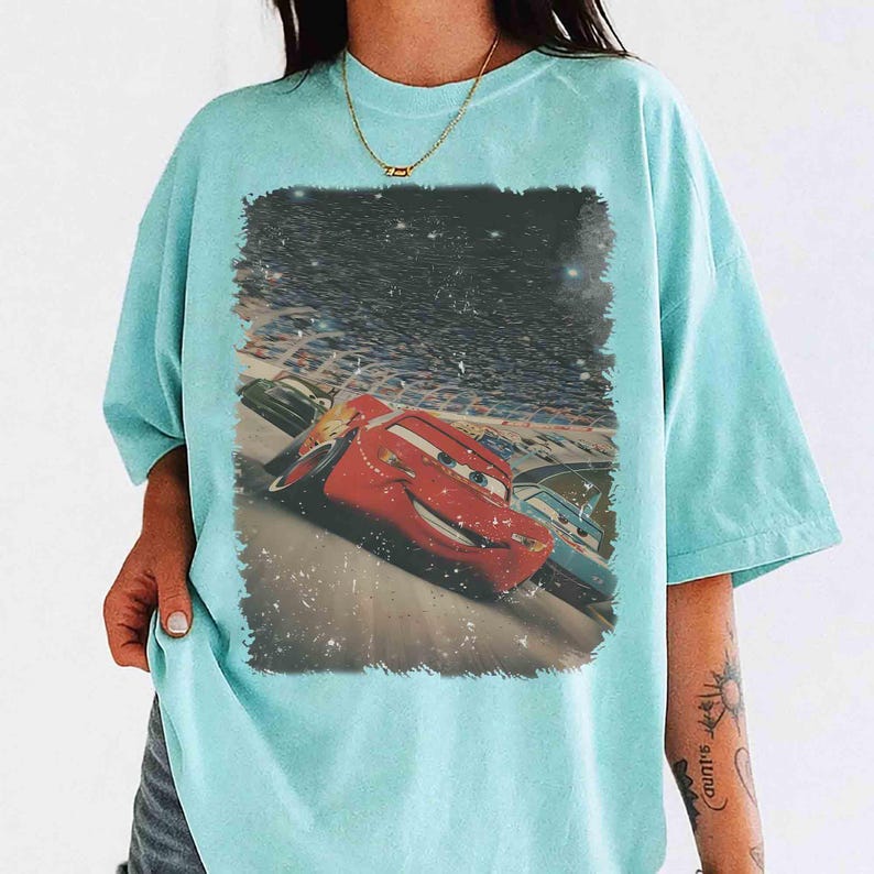 Puede incluir: Una camiseta azul claro con un gr&aacute;fico de Rayo McQueen de la pel&iacute;cula Cars corriendo en una pista. El gr&aacute;fico est&aacute; enmarcado en un rect&aacute;ngulo con un aspecto vintage y desgastado. La camiseta tiene mangas cortas y cuello redondo.