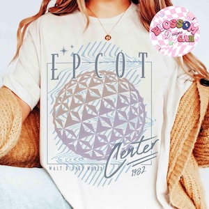 May include: Cream-colored t-shirt featuring a graphic of the EPCOT Center sphere with the text "EPCOT Center" and "Walt Disney World 1982". The design includes blue and purple tones.