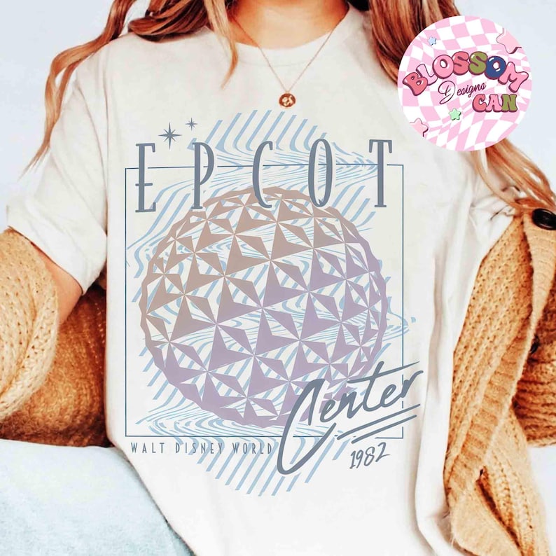 May include: Cream-colored t-shirt featuring a graphic of the EPCOT Center sphere with the text "EPCOT Center" and "Walt Disney World 1982". The design includes blue and purple tones.