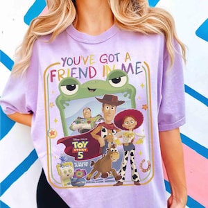 May include: A light purple t-shirt featuring a graphic print with the text "YOU'VE GOT A FRIEND IN ME" and "TOY STORY 5 IN THEATERS JUNE 19". The design includes illustrations of Buzz Lightyear, Woody, Jessie, Bullseye, and a green frog frame.