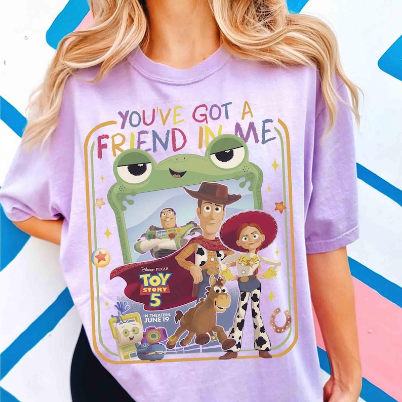 May include: A light purple t-shirt featuring a graphic print with the text "YOU'VE GOT A FRIEND IN ME" and "TOY STORY 5 IN THEATERS JUNE 19". The design includes illustrations of Buzz Lightyear, Woody, Jessie, Bullseye, and a green frog frame.