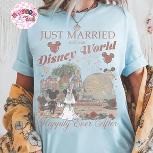 May include: Light blue t-shirt with the words "Just Married Disney World" and "Happily Ever After" printed on the front. The design includes Mickey and Minnie Mouse, balloons, and iconic Disney World landmarks. The shirt is a short-sleeved crew neck.