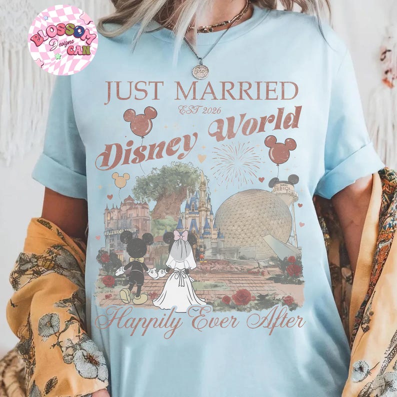 May include: Light blue t-shirt with the words "Just Married Disney World" and "Happily Ever After" printed on the front. The design includes Mickey and Minnie Mouse, balloons, and iconic Disney World landmarks. The shirt is a short-sleeved crew neck.