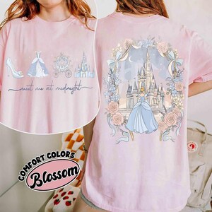 May include: Pink t-shirt with a Cinderella theme. The front features a high heel, dress, carriage, and castle with the text "meet me at midnight." The back displays Cinderella in a blue dress in front of a castle, surrounded by flowers and ribbons.