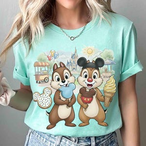 May include: A light teal t-shirt featuring Chip and Dale holding treats in front of a cartoon scene with a castle, sun, and food carts. Chip holds a blue ice cream, and Dale holds a Mickey Mouse ice cream. A Mickey Mouse cookie is also visible.