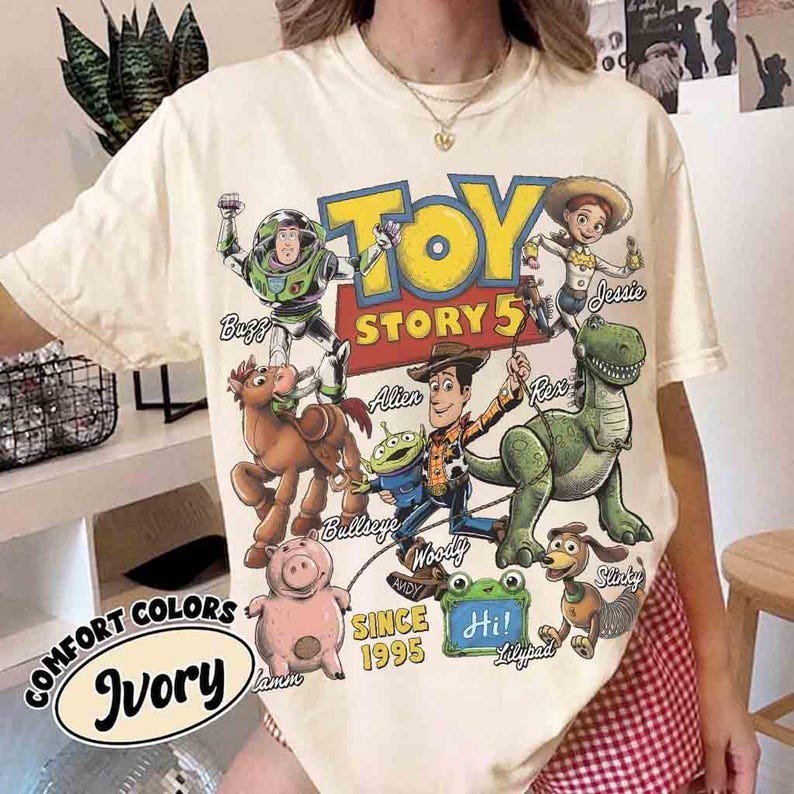 May include: An ivory short-sleeve t-shirt featuring a colorful graphic print of Toy Story characters including Buzz Lightyear, Woody, Jessie, Bullseye, Rex, Hamm, Slinky Dog, and an Alien. The text reads Toy Story 5, Andy, Since 1995, and Hi.