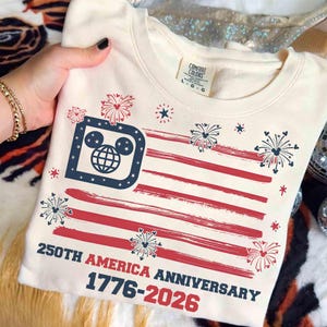 May include: Cream-colored sweatshirt featuring a stylized American flag design with red stripes and a blue square with a Mickey Mouse head and globe. The text reads "250th America Anniversary 1776-2026". Fireworks and stars complete the patriotic theme.