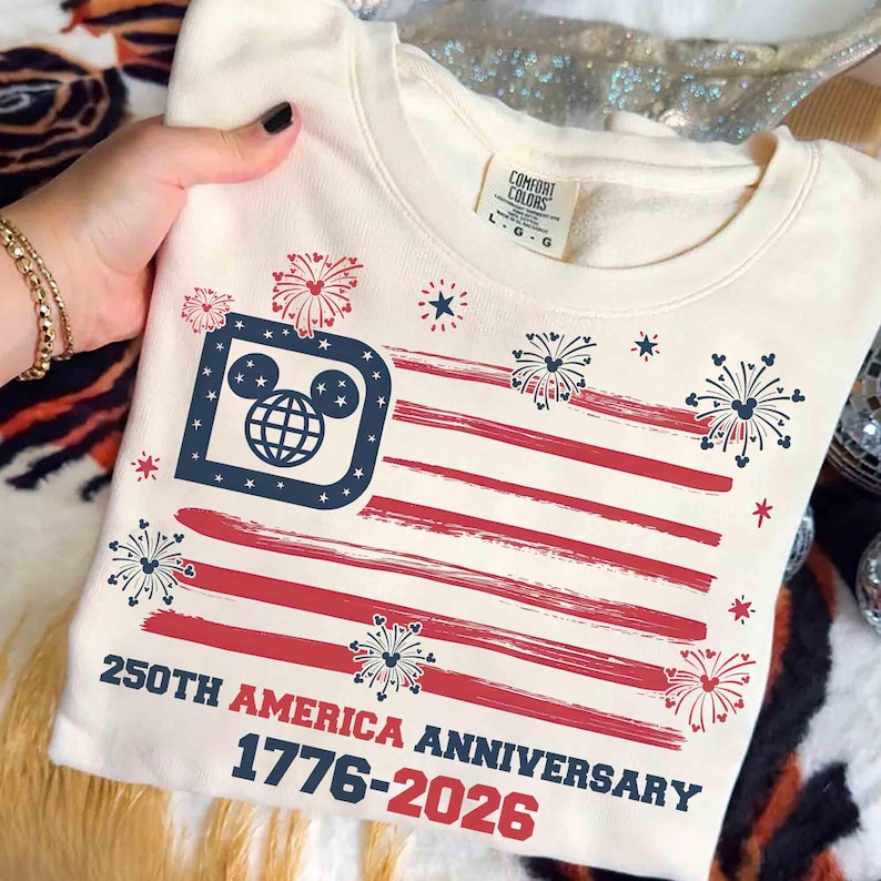 May include: Cream-colored sweatshirt featuring a stylized American flag design with red stripes and a blue square with a Mickey Mouse head and globe. The text reads "250th America Anniversary 1776-2026". Fireworks and stars complete the patriotic theme.
