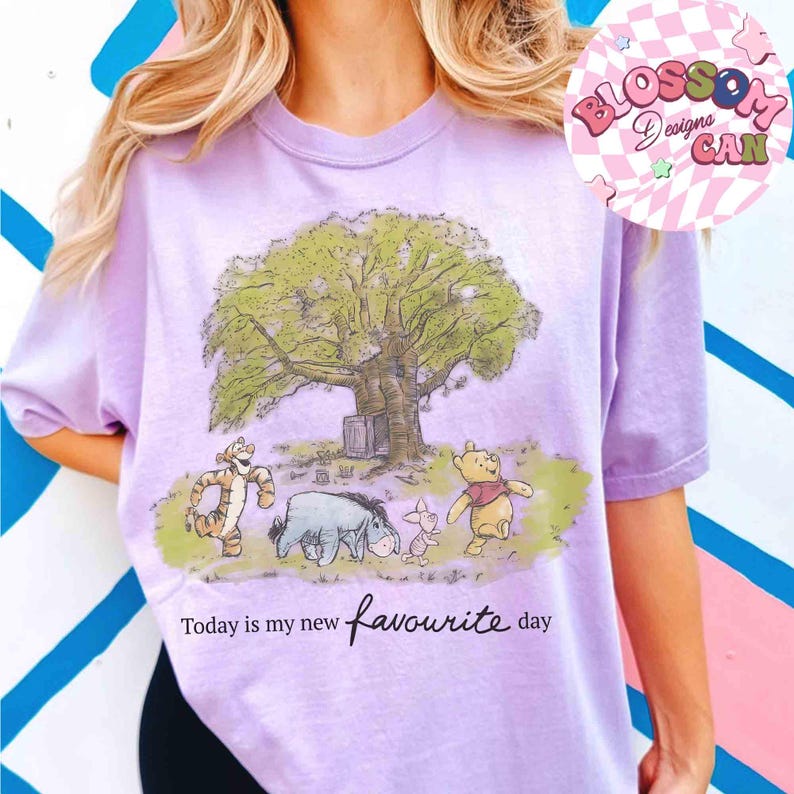 K&ouml;nnte beinhalten: Ein lilafarbenes T-Shirt mit einer Illustration von Winnie Puuh und seinen Freunden unter einem Baum. Das Design zeigt Tiger, I-Aah, Ferkel und Puuh. Der Text "Today is my new favourite day" ist darunter gedruckt.