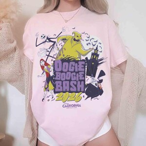 May include: A light pink t-shirt featuring characters from The Nightmare Before Christmas. The shirt has the text "Oogie Boogie Bash 2026" and "Disney California Adventure". The design includes Jack Skellington, Sally, Oogie Boogie, and Zero.