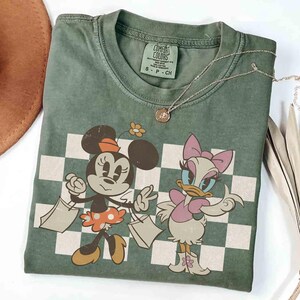 May include: Green Comfort Colors t-shirt featuring Minnie Mouse and Daisy Duck with shopping bags in front of a checkered background. A silver necklace with a small pendant is on the shirt. The shirt has a small tag that says "S-P-CH".