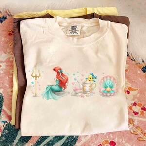 May include: Cream-colored t-shirt featuring a colorful illustration of a mermaid, a fish, and a seashell. The mermaid has red hair and a teal tail. The shirt is folded on top of two other shirts, one brown and one yellow.