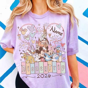 K&ouml;nnte beinhalten: Ein helllila T-Shirt mit einem Disney-Motiv. Das Design zeigt Micky Maus, Minnie Maus und Donald Duck vor einem Schloss. Der Text "Disney Trip 2026" steht unter den Figuren, und "Name" steht dar&uuml;ber. Das T-Shirt hat kurze &Auml;rmel.
