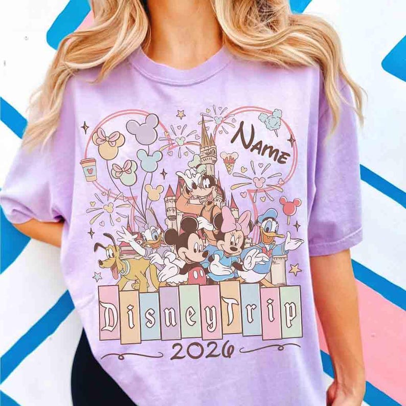 K&ouml;nnte beinhalten: Ein helllila T-Shirt mit einem Disney-Motiv. Das Design zeigt Micky Maus, Minnie Maus und Donald Duck vor einem Schloss. Der Text "Disney Trip 2026" steht unter den Figuren, und "Name" steht dar&uuml;ber. Das T-Shirt hat kurze &Auml;rmel.