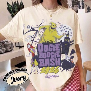 May include: Ivory-colored t-shirt featuring a graphic design with characters from The Nightmare Before Christmas. The design includes the text "Oogie Boogie Bash 2026" and "Disney California Adventure". The shirt is part of the Comfort Colors collection.