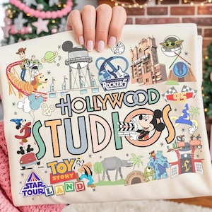Può includere: Una maglietta color crema con un design grafico colorato del parco a tema Hollywood Studios. Il design include personaggi dei cartoni animati, attrazioni del parco e le parole "Hollywood Studios" a grandi lettere. La maglietta è tenuta da una persona.